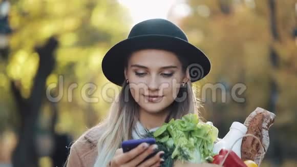 快乐的年轻美丽的女人穿着时尚的外套在公园里散步拿着一包产品用智能手机视频的预览图