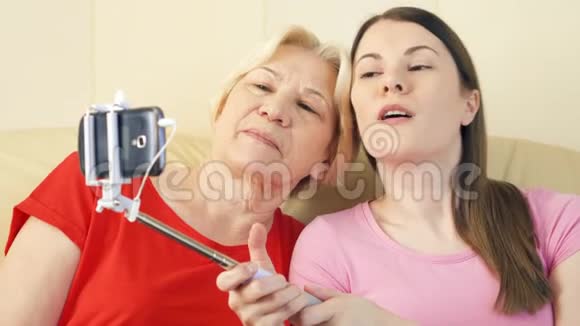 母亲和女儿坐在家里的沙发上用智能手机和自拍棒制作自拍视频的预览图