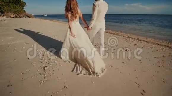 新娘和新郎赤脚走在蓝色海洋旁边的沙滩上他们牵着手一起快乐视频的预览图
