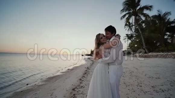 新娘和新郎在海边日落时在美丽的热带海滩上亲吻棕榈树浪漫的婚姻视频的预览图
