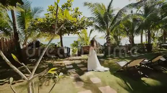 一位穿着白色长裙的美丽新娘沿着棕榈树之间的小径走向新郎庄严时刻这就是视频的预览图