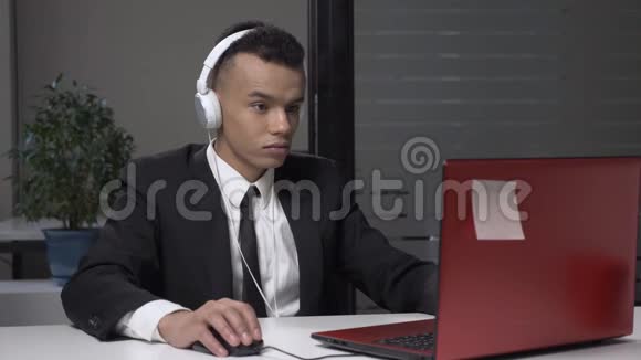 年轻成功的非洲商人穿着西装戴着耳机在电脑上工作在工作中玩电脑游戏视频的预览图