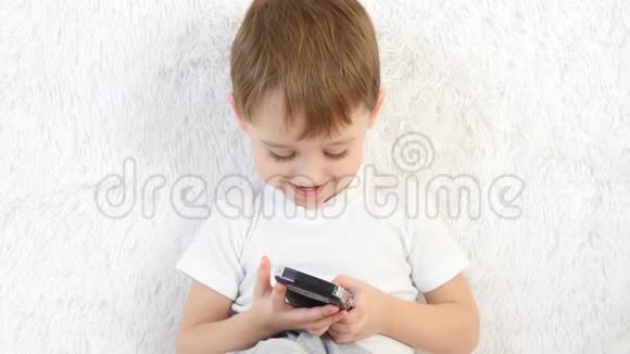 一个孩子正坐在白色沙发上玩着智能手机特写孩子对游戏很满意微笑着视频的预览图