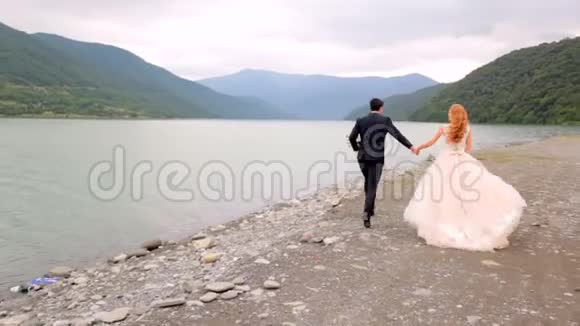 新婚快乐的新娘和新郎在美丽的群山背景下沿着河岸奔跑格鲁吉亚视频的预览图