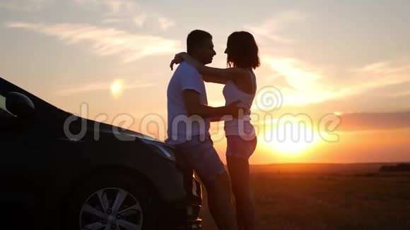 幸福夫妇的剪影停留在新车的天空下日落时分美丽的年轻夫妇站在汽车附近视频的预览图