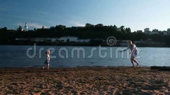 日落时分美丽的金发母亲和女儿在河岸上朝对方跑去视频的预览图