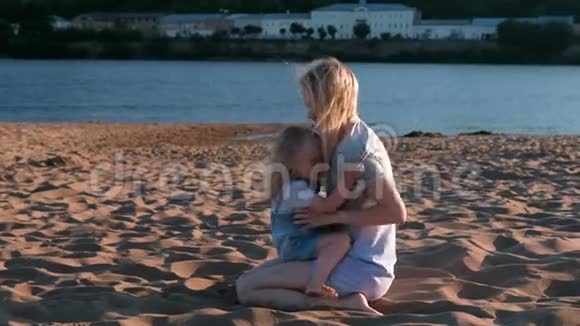 美丽快乐的金发妈妈和女儿拥抱和说话坐在海滩上日落视频的预览图
