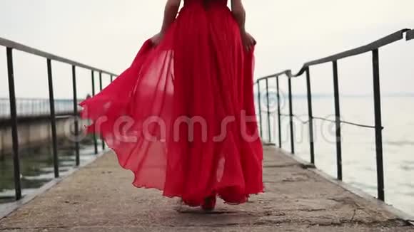 穿着猩红裙子的快乐美女穿着红色高跟鞋在码头的海边漫步视频的预览图