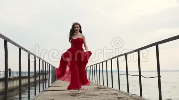 一位身穿猩红连衣裙卷发的年轻美女沿着码头沿着大海奔跑视频的预览图