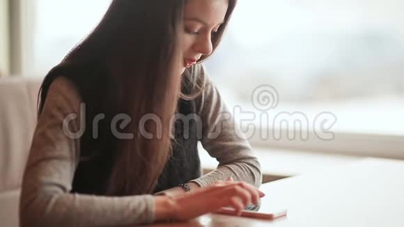 一个年轻漂亮的女孩手里拿着一部手机坐在咖啡馆的桌子旁休息一下视频的预览图