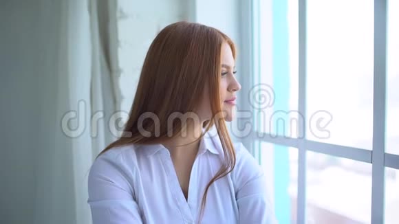 一个微笑的红头发女孩坐在家靠近窗户的肖像一个微笑的女学生的肖像女人微笑视频的预览图