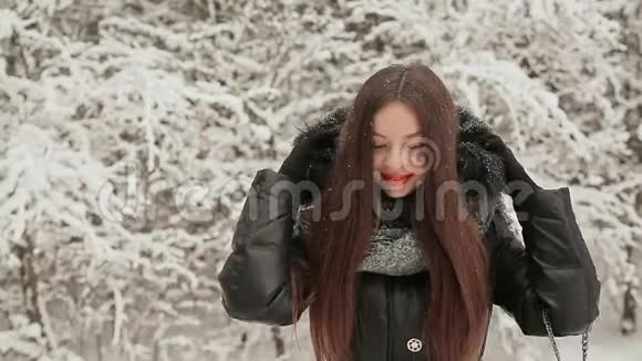 在白雪覆盖的杉树的背景下一个年轻漂亮的女孩穿着暖冬的衣服带着一个引擎盖不被遮挡视频的预览图