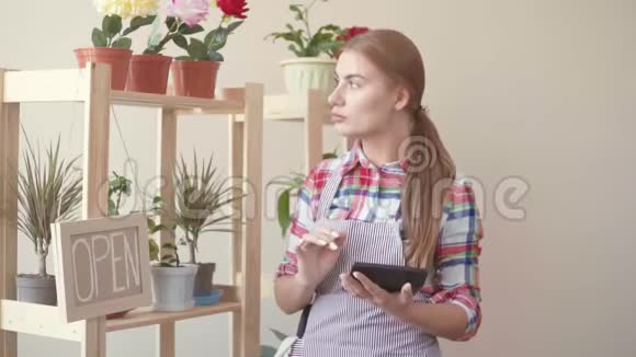 小生意一个穿着格子衬衫和条纹围裙的女孩站在那里手里拿着一块数字平板电脑并计划着花卉师视频的预览图
