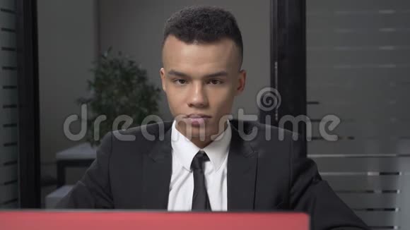 年轻成功的非洲商人坐在办公室用笔记本电脑工作看着相机微笑着画像视频的预览图