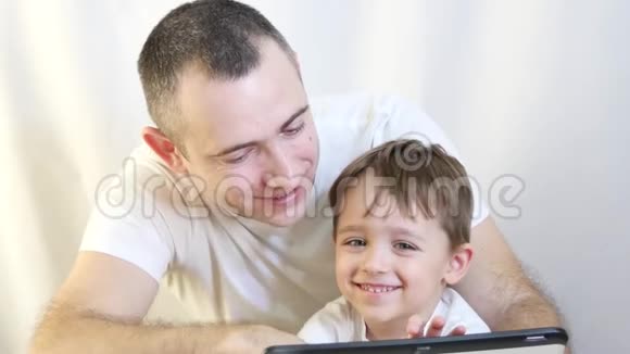 父亲和儿子坐在桌子上在平板电脑上玩耍在白色的背景上被隔离幸福家庭的概念视频的预览图