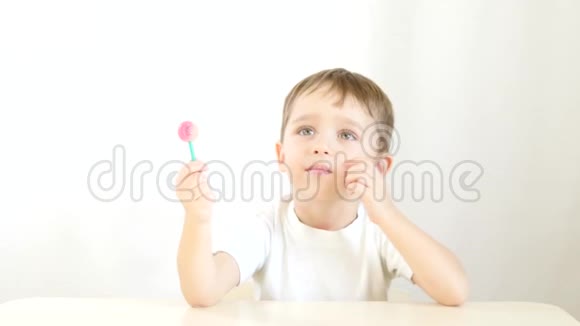 快乐的孩子高兴地舔着棒棒糖坐在白色背景的桌子旁视频的预览图