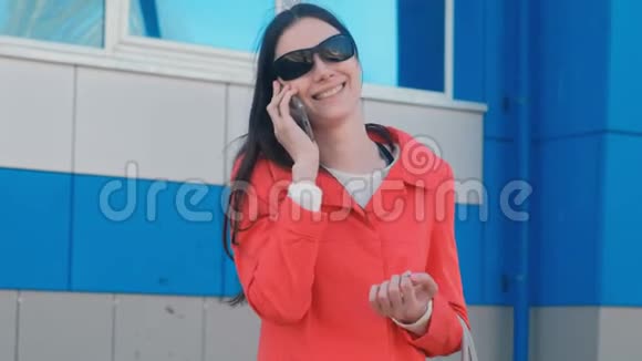 一幅年轻的黑发女人戴着太阳镜和红色外套的肖像在蓝色建筑旁边的电话里说话视频的预览图