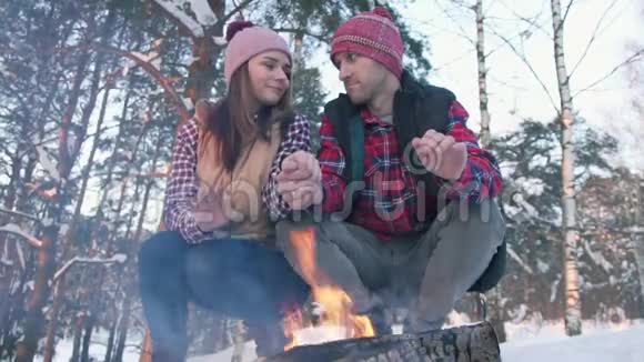 一对穿着红色衬衫的年轻人在树林里温暖他们的手在火边冬季野餐视频的预览图