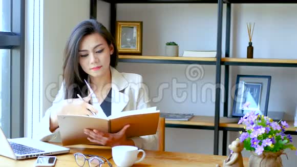 4K镜头美丽的亚洲商务女性坐在电脑笔记本电脑前阅读并翻翻笔记簿页面和思考视频的预览图
