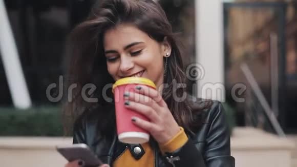年轻快乐的女人使用智能手机喝咖啡去户外沿着城市的街道走慢动作视频的预览图