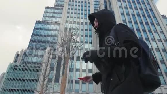 穿着黑色夹克和背包的男人用手机在城市大街上行走视频的预览图