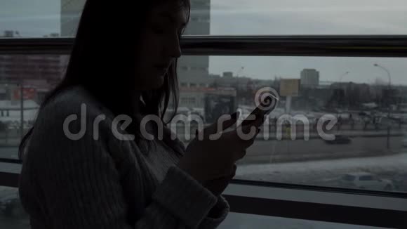 年轻的女孩穿着灰色毛衣在特大城市背景的窗户旁的咖啡馆里带着电话视频的预览图