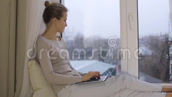 漂亮的女人在笔记本电脑上打字坐在窗台上视频的预览图