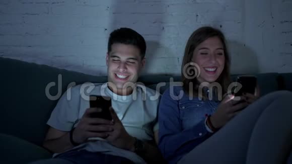 年轻的快乐和浪漫的一对20多岁的夫妇一起使用手机享受坐在家里的沙发上笑着玩视频的预览图