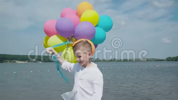 快乐的小男孩带着五颜六色的气球在湖岸散步4K视频的预览图
