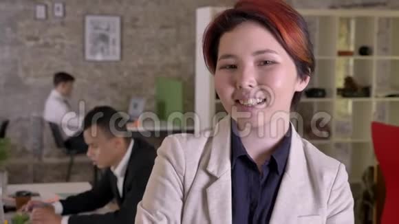年轻的商业亚洲女人在现代办公室里看着镜头微笑着黑人和高加索男人在后面工作视频的预览图