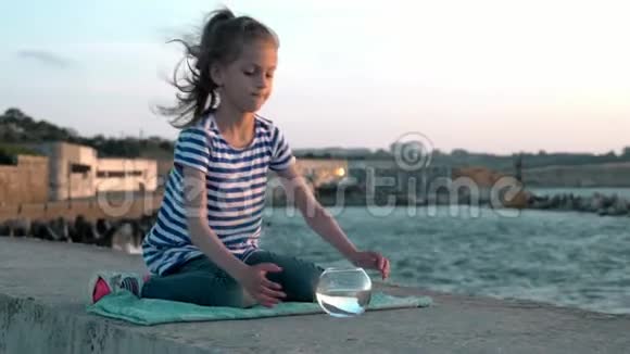 小女孩坐在背景城市码头的海景日落吃小鱼缸看小鱼概念再现视频的预览图