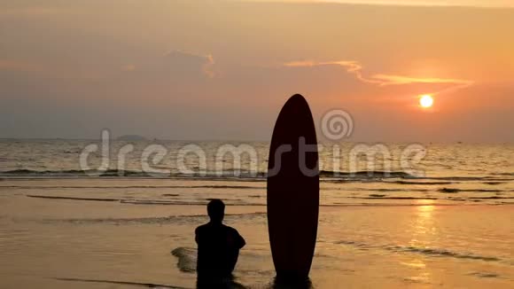 4K冲浪者坐在海边的剪影在热带海滩日落时有长长的冲浪板视频的预览图