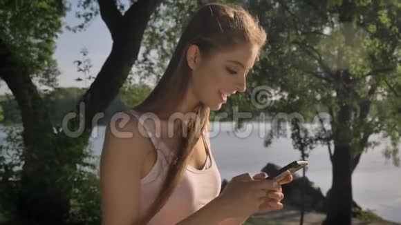 年轻漂亮的女人在手机上发短信看着镜头微笑着停在河边的背景附近视频的预览图