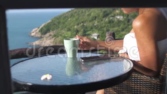 年轻女子裹着毛巾在阳台上喝咖啡享受海景并使用手机3840x2160视频的预览图