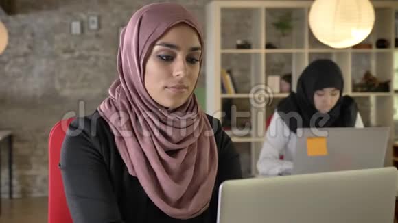 年轻的穆斯林妇女工作严肃两个女人戴着头巾坐着在现代办公室的笔记本电脑上打字视频的预览图