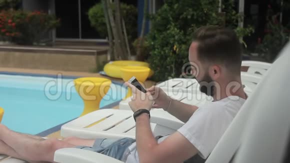 年轻人正坐在游泳池附近的躺椅上在他的智能手机上打字视频的预览图