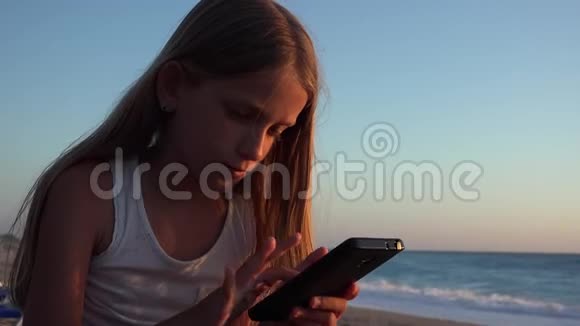 儿童玩智能手机孩子在日落海滩女孩在海滨使用平板电脑视频的预览图