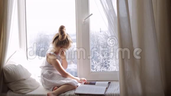 可爱的小女孩一本书坐在窗台上窗外的冬天视频的预览图