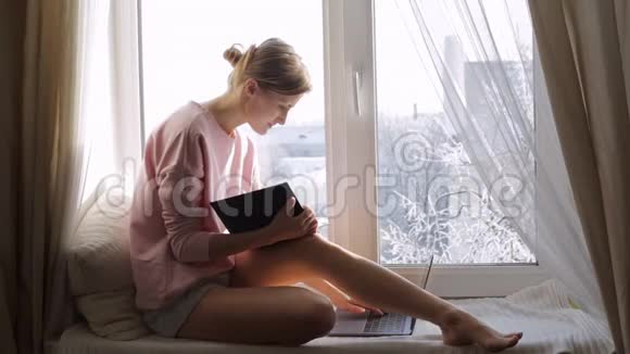 一个拿着记事本的年轻女人坐在窗台上用电脑视频的预览图