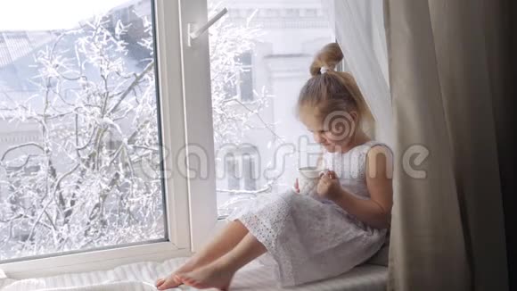 可爱的小女孩一杯茶坐在窗台上带着杯子的女孩透过窗户看冬天晴朗视频的预览图
