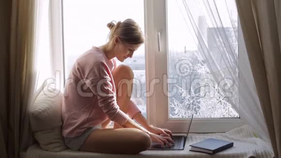 年轻女子手提电脑坐在窗台上然后她透过窗户看视频的预览图