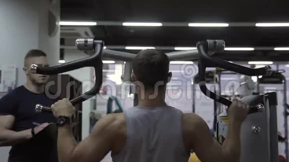 年轻的运动员正在健身房里拉体重机他的男性教练正在控制他视频的预览图