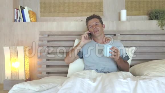 年轻漂亮的30岁男人蓝眼睛在家里快乐放松在床上用手机笑着说话视频的预览图