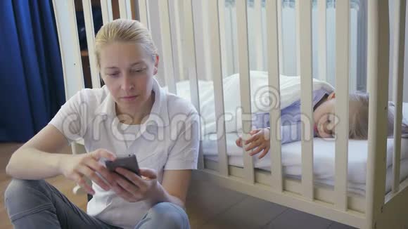 一个手里拿着智能手机的女人坐在婴儿床上熟睡的小婴儿旁边视频的预览图