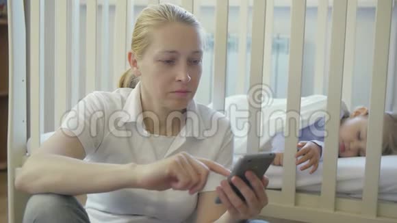一个手里拿着智能手机的女人坐在婴儿床上熟睡的小婴儿旁边视频的预览图