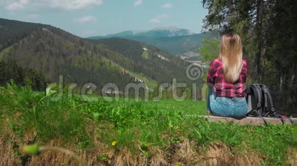一个年轻的高加索女人带着背包坐在山顶上的一根木头上戴着一顶帽子的概念视频的预览图