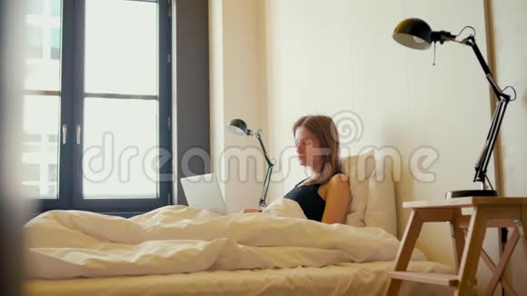 年轻的商务女性正在舒适的家庭客房里从床上拿出笔记本电脑视频的预览图