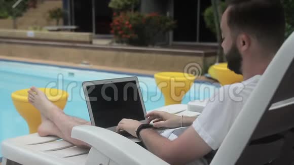 坐在躺椅上的池边长胡子的人很快在笔记本电脑上打字视频的预览图