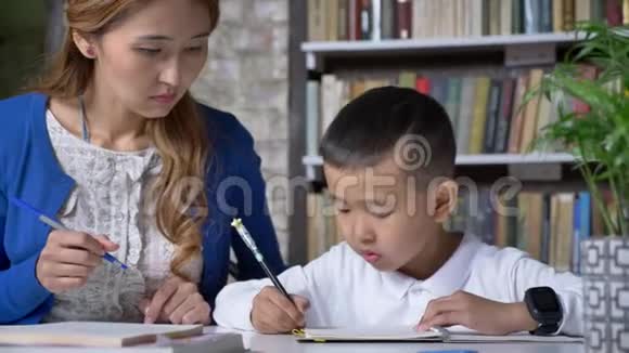 亚洲小男孩和年轻的母亲一起做家庭作业女人帮助孩子坐在桌子后面书架背景视频的预览图
