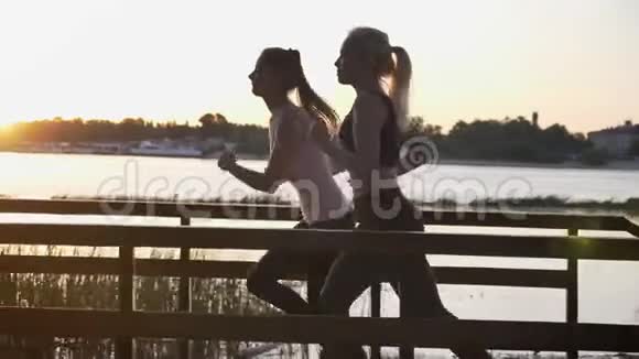 日落时分两位年轻漂亮的女士在河边公园跑步减肥健身模特慢跑美丽的景色视频的预览图